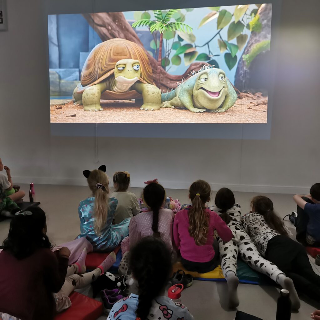 Children sitting on mats watching an animated movie on a large screen
