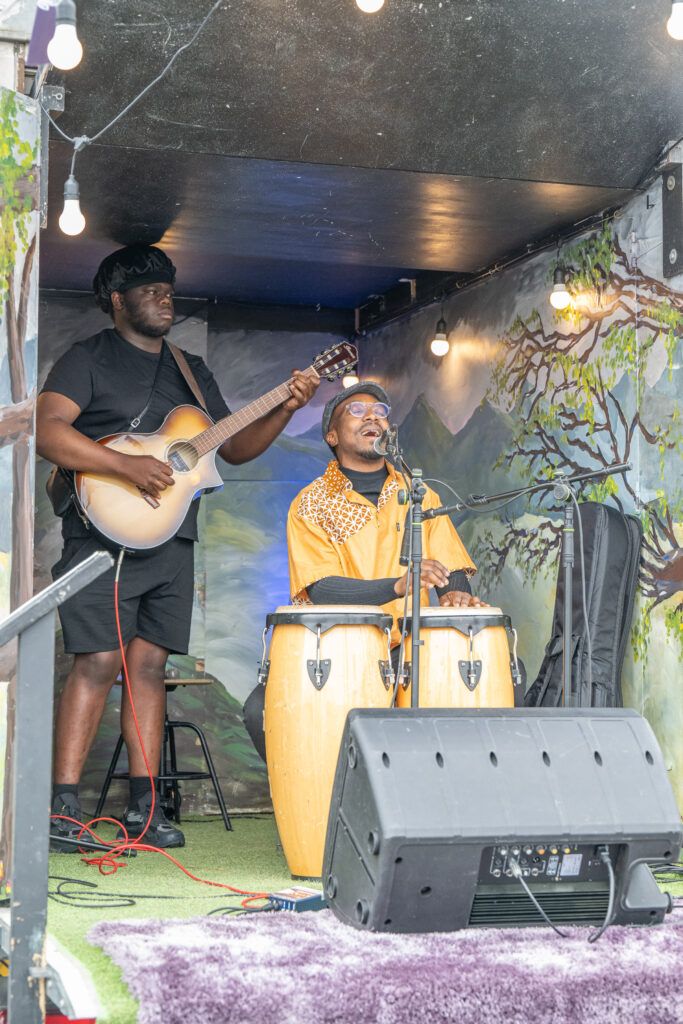 Two musicians performing with guitar and drums on a small stage at a festival