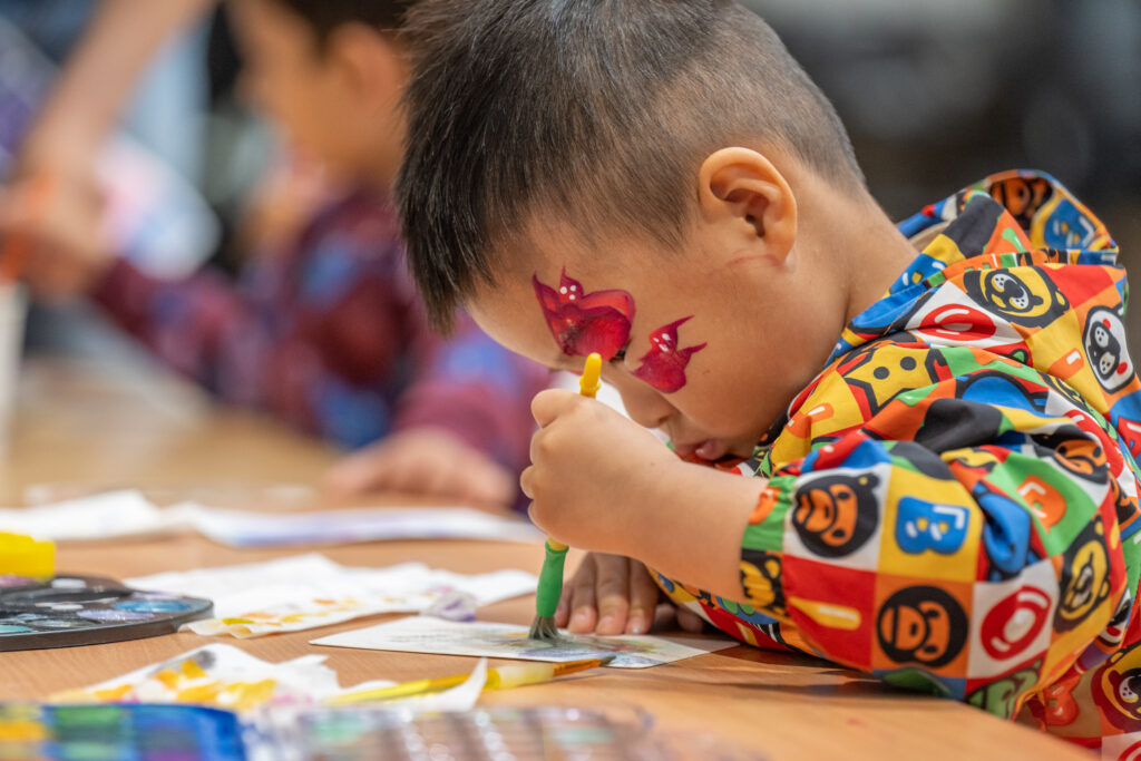 Child with butterfly face paint painting at an arts and crafts table during a festival